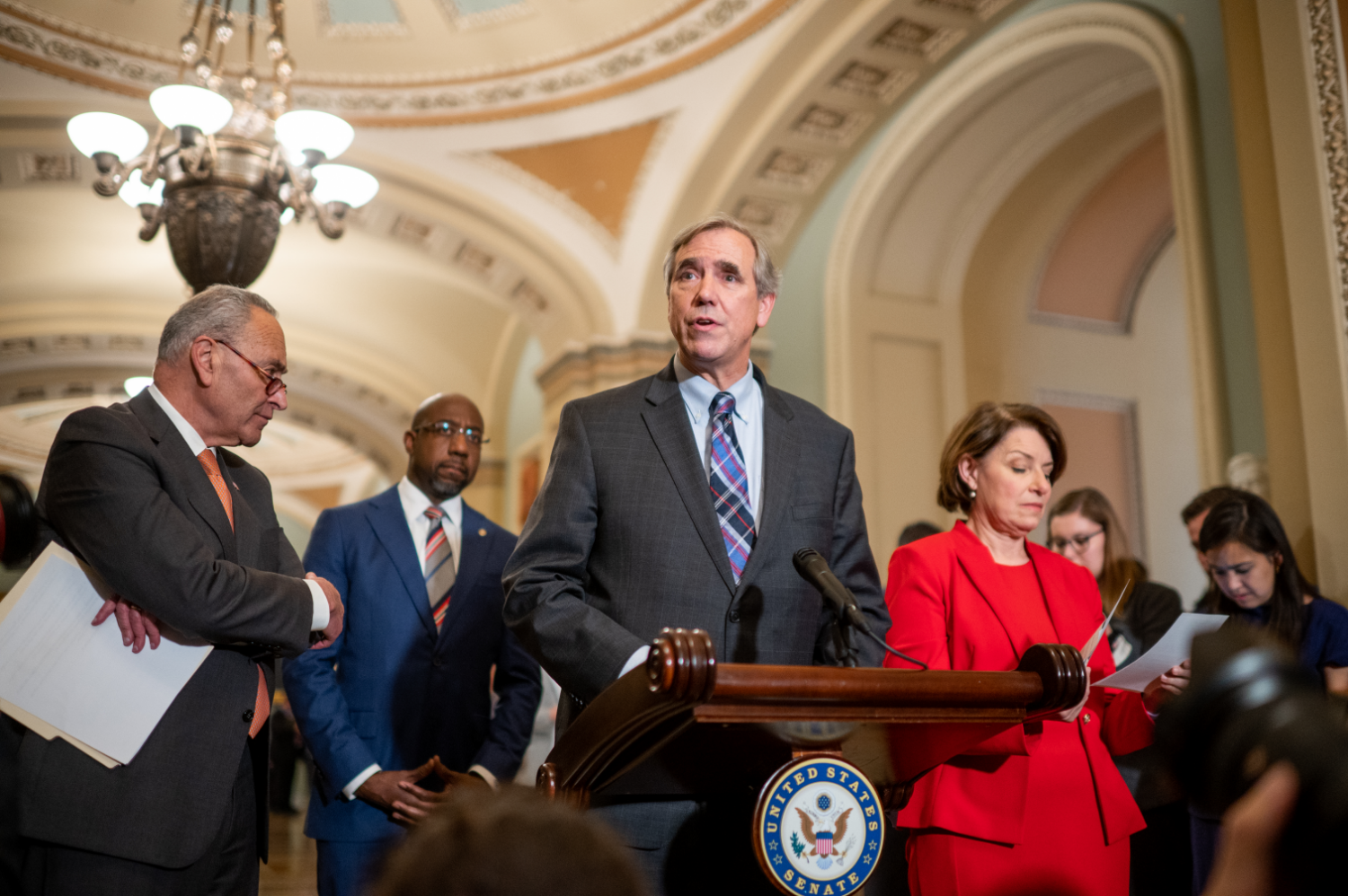 Senator Jeff Merkley speaking at a podium with the seal of the U.S. Senate on the front.